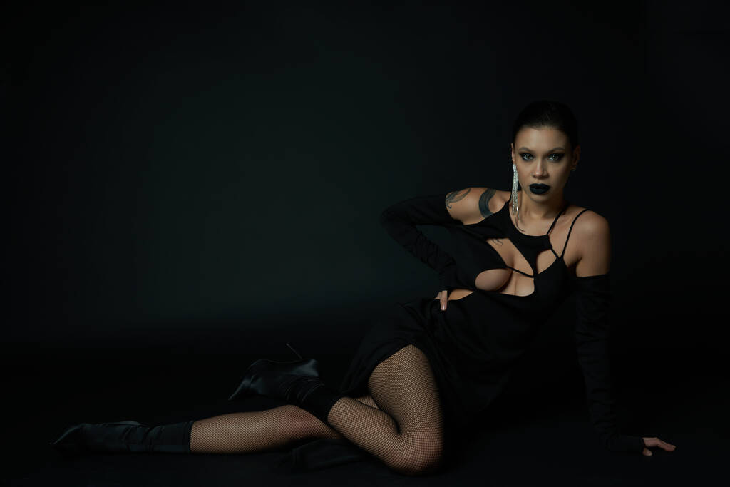 seductora mujer tatuada en vestido de Halloween sentado y mirando a la cámara en negro, siniestra belleza - Foto, Imagen