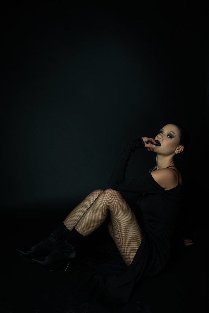 sensual woman in sexy halloween dress and dark makeup looking at camera while sitting on black - Photo, Image