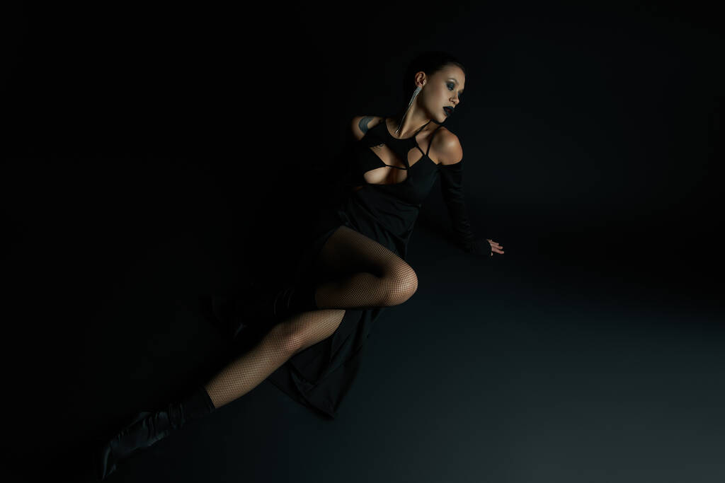 τατουάζ δαίμονας-εμπνευσμένη γυναίκα σε κομψό αποκριάτικο φόρεμα κάθεται σε μαύρο φόντο, πλήρους μήκους - Φωτογραφία, εικόνα