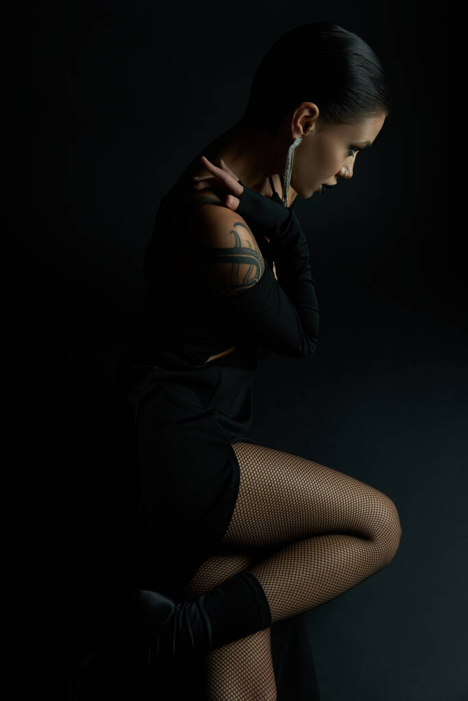 boční pohled na svůdné tetované ženy v sexy šaty a punčocháče sedí na černé, tmavá krása - Fotografie, Obrázek