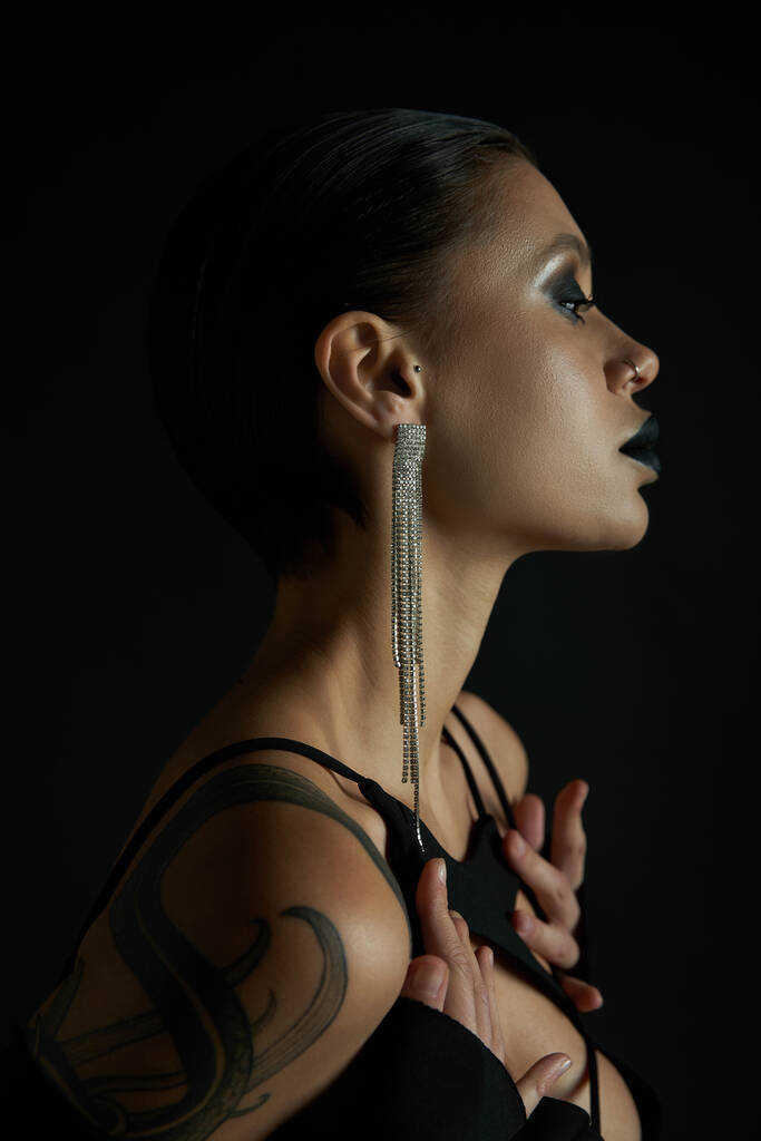 profilo di sensuale donna tatuata con trucco scuro e orecchino lucido su nero, concetto di Halloween - Foto, immagini