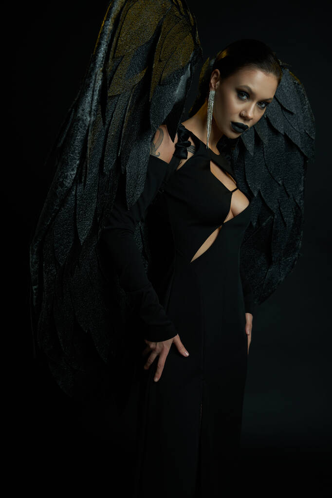 сексуальная татуированная женщина в костюме падшего ангела на Хэллоуин с темными крыльями, смотрящая в камеру на черном - Фото, изображение