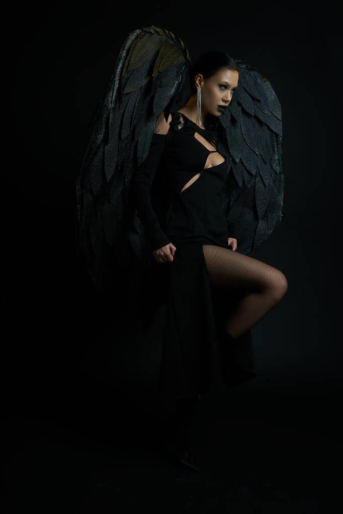uwodzicielska kobieta w ciemnym makijażu i kostiumie z demonicznymi skrzydłami odwracając wzrok na czarno, Halloween - Zdjęcie, obraz