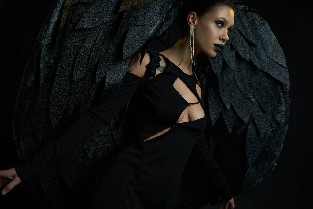 Cadılar bayramı kostümlü büyüleyici bir kadın. Kara melek kanatlarıyla kara arka plana bakıyor. - Fotoğraf, Görsel
