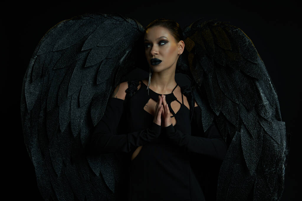 mujer tatuada en traje de ángel oscuro de pie con las manos de oración y mirando hacia otro lado en negro - Foto, imagen