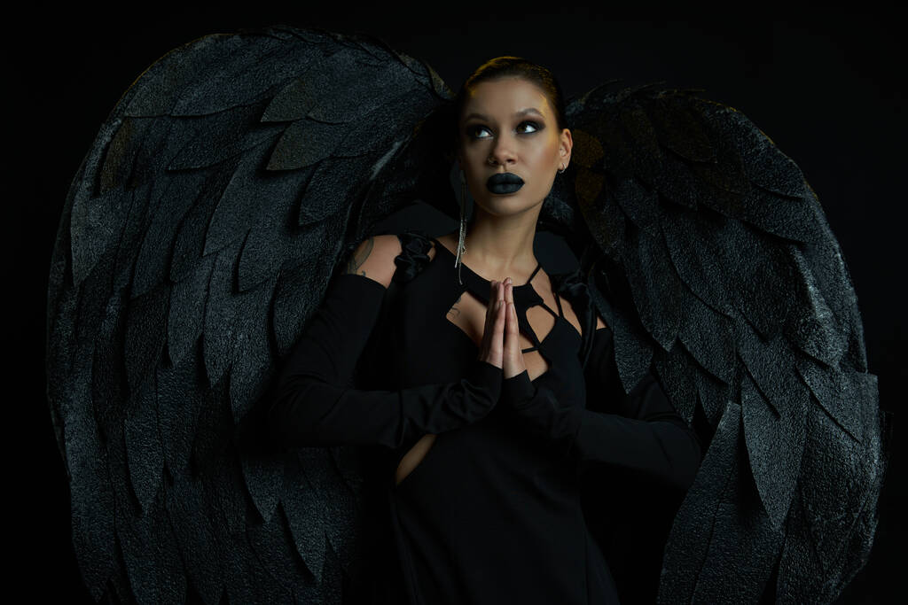 sexy donna in costume di scuro angelo caduto in piedi con le mani in preghiera e guardando lontano su nero - Foto, immagini