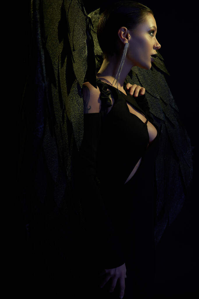 δαιμονική γοητεία, πλαϊνή άποψη της σέξι γυναίκα στο αποκριάτικο κοστούμι του σκοτεινού δαίμονα με φτερά στο μαύρο - Φωτογραφία, εικόνα