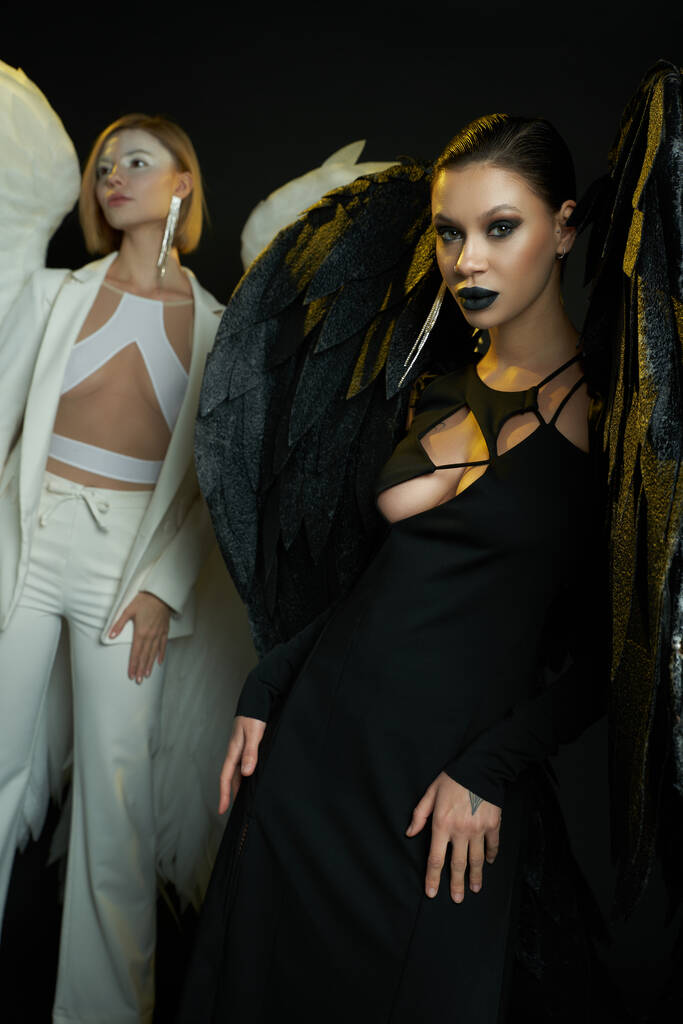 demonische vrouw kijken naar camera in de buurt van engel op zwarte achtergrond, vrouwen in kostuums van gevleugelde wezens - Foto, afbeelding