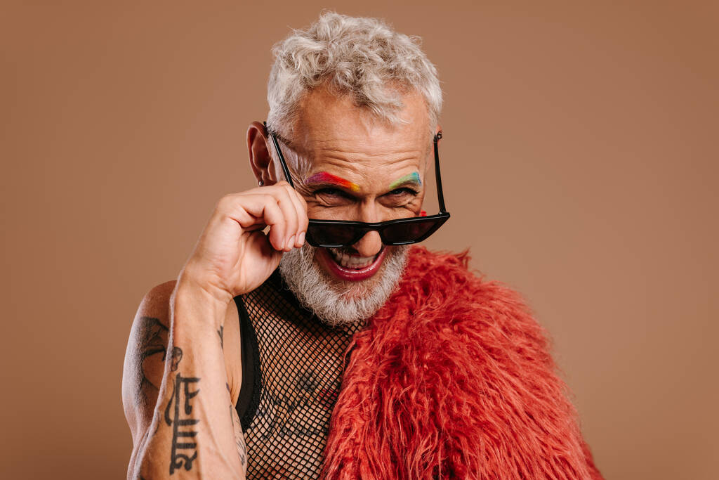 Стильный зрелый гей с цветными бровями, регулирующий очки и улыбающийся на коричневом фоне - Фото, изображение