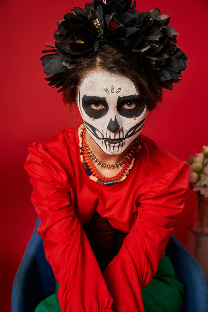 mujer en aterrador maquillaje de cráneo de azúcar y cuentas de colores mirando a la cámara en sillón en rojo - Foto, imagen