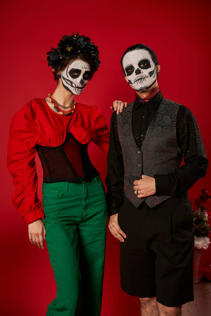 elegant paar in dia de los muertos make-up en feestelijke kleding kijken naar camera in de buurt van bloemen op rood - Foto, afbeelding