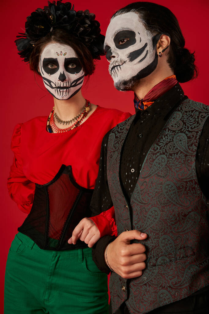 Kafatası makyajlı kadın ve siyah çelenk kırmızı giyen adamın yanındaki kameraya bakıyor, dia de los muertos çifti - Fotoğraf, Görsel