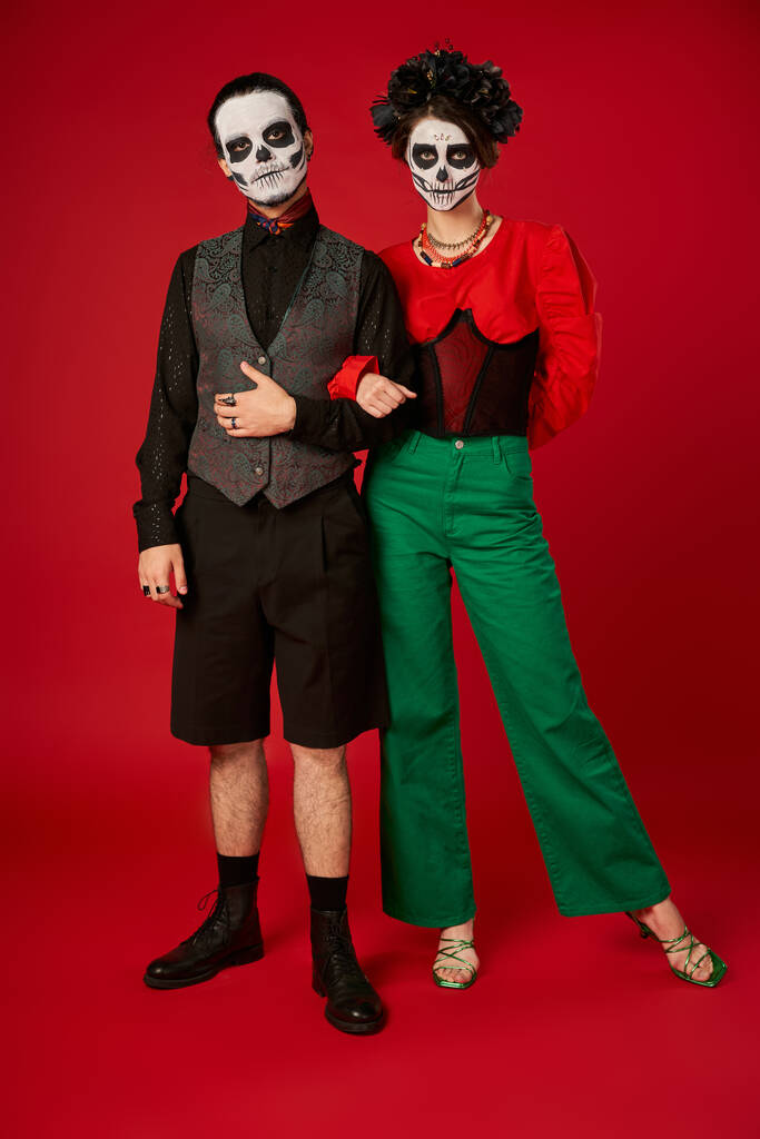full length of couple in trendy clothes and skeleton makeup on red, dia de los muertos celebration - Photo, Image