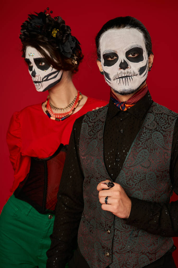 mężczyzna w makijaż czaszki patrząc w aparat w pobliżu kobieta w czarny wieniec, dia de los muertos para na czerwony - Zdjęcie, obraz