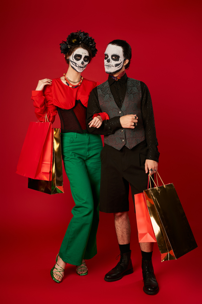full length of couple in dia de los muertos μακιγιάζ στέκεται με τσάντες για ψώνια σε κόκκινο, εορταστική πώληση - Φωτογραφία, εικόνα