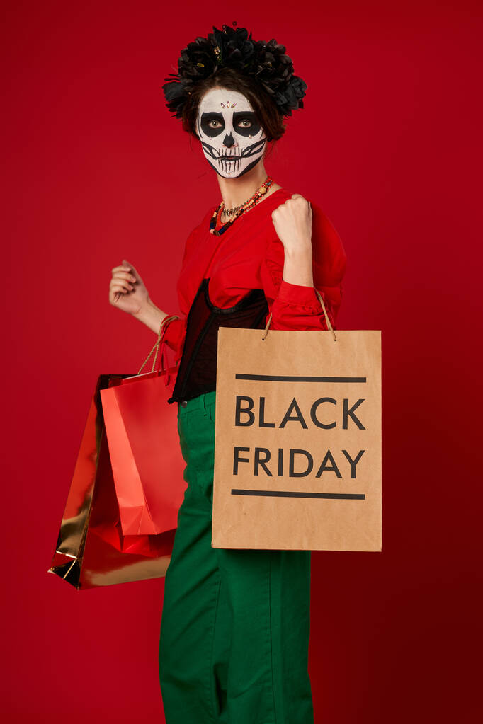 stylish woman in dia de los muertos makeup standing with shopping bags on red, black friday sale - Photo, Image