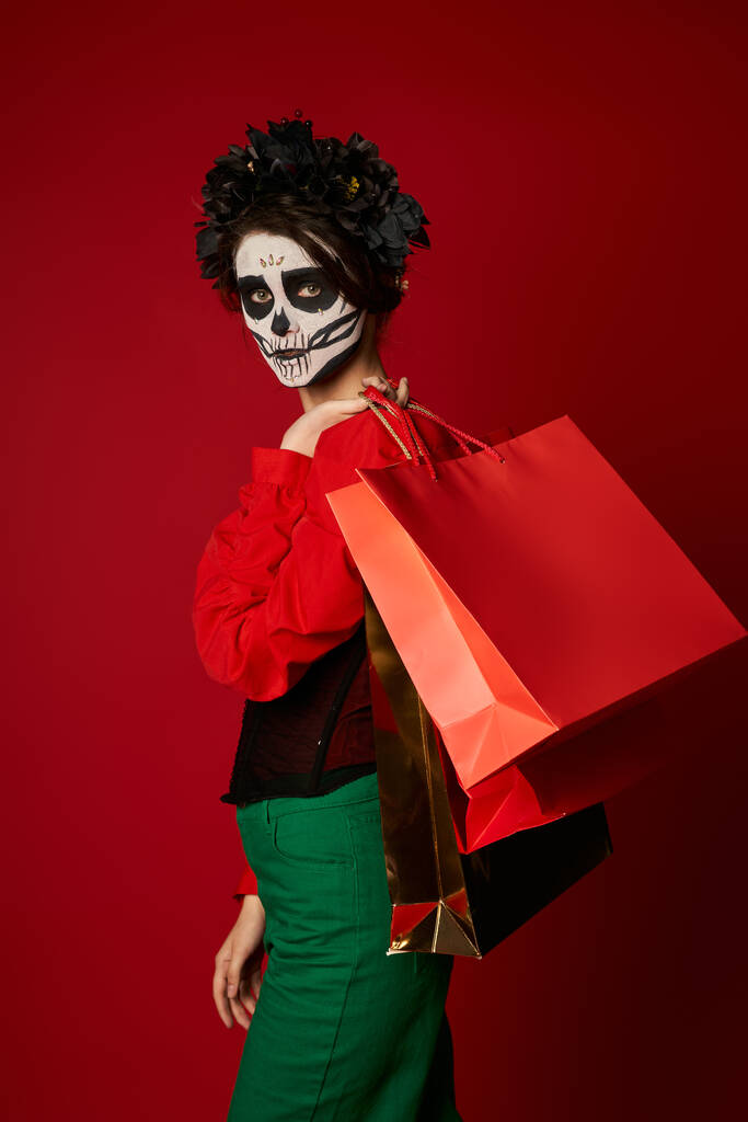 mulher elegante em coroa preta e maquiagem catrina segurando sacos de compras e olhando para a câmera no vermelho - Foto, Imagem