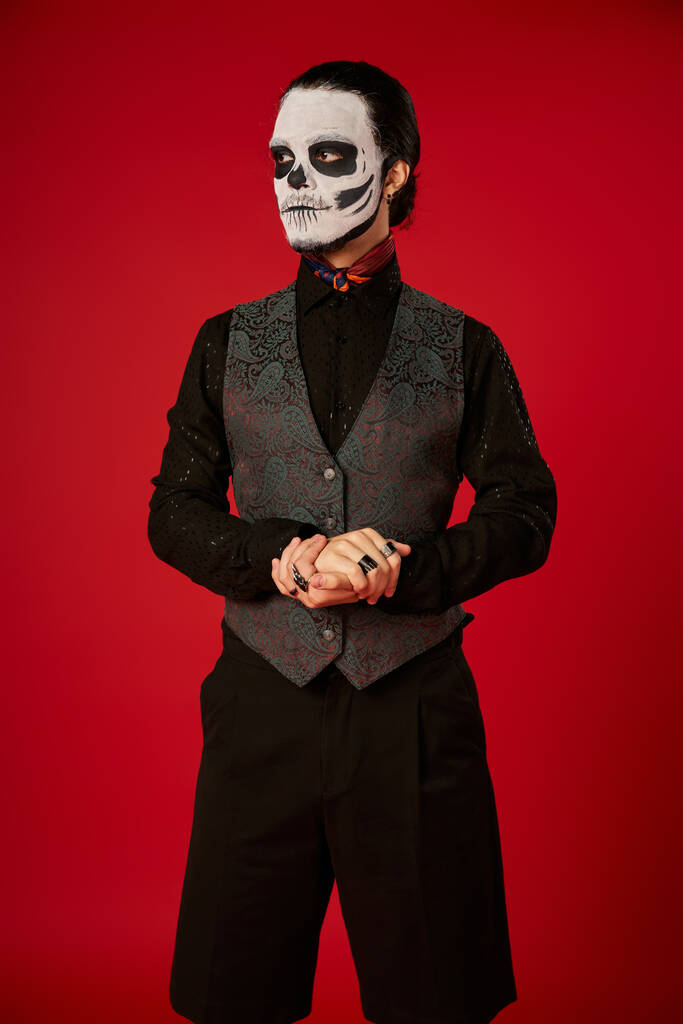homme en tenue de fête et maquillage du crâne de sucre regardant loin sur rouge, dia de los muertos célébration - Photo, image