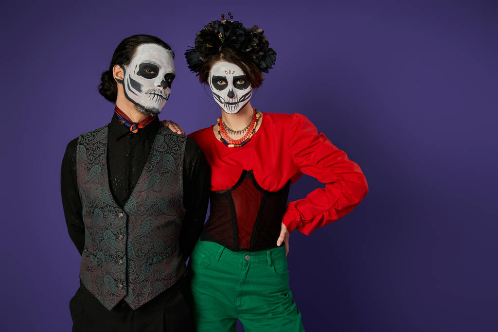 couple élégant dans le maquillage squelette traditionnel dia de los muertos regardant la caméra sur fond bleu - Photo, image