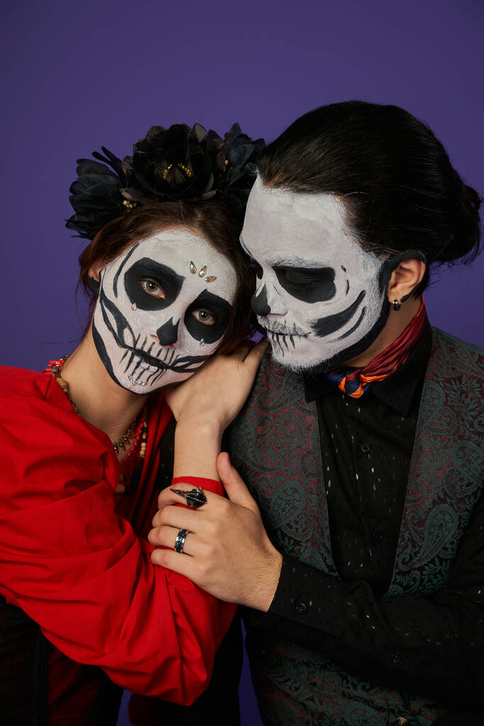 woman in skull makeup and black wreath leaning on shoulder of man and looking at camera on blue - Photo, Image