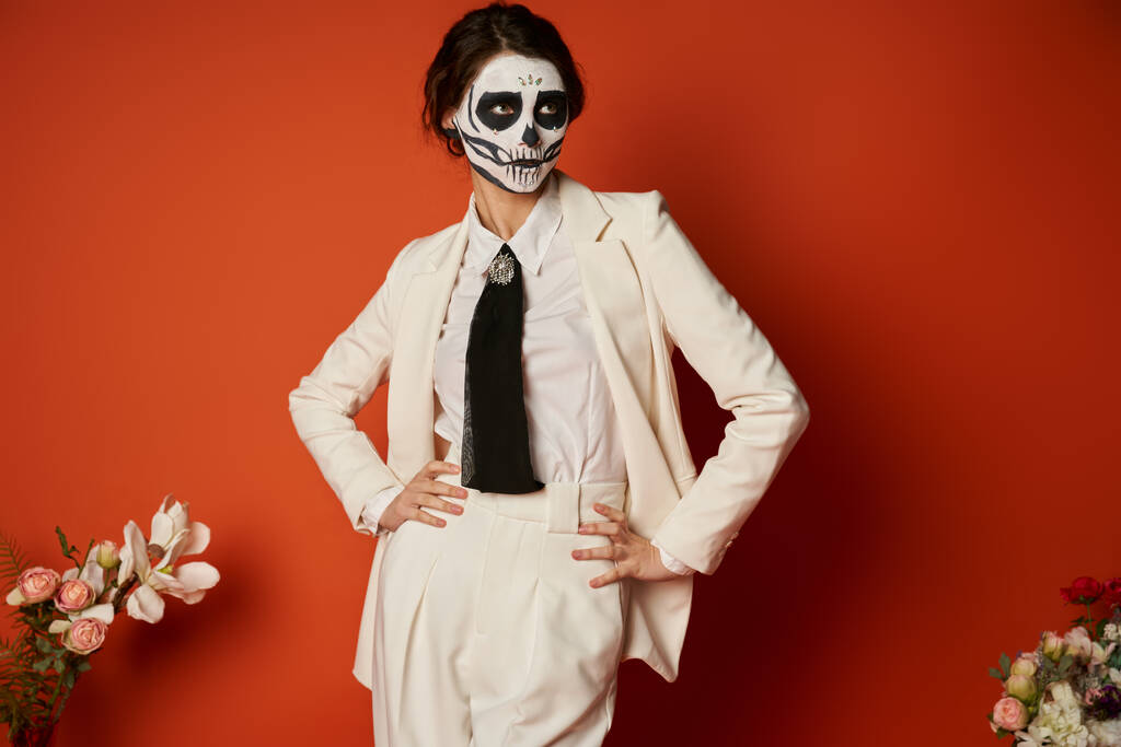 женщина в скелете макияж и белый костюм позирует с руками на бедрах рядом с цветами на красный, День мертвых - Фото, изображение