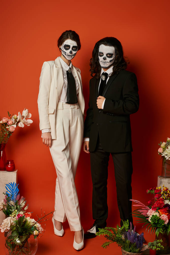 пара в сахарном черепе макияж и элегантные костюмы рядом с dia de los muertos алтарь с цветами на красном - Фото, изображение