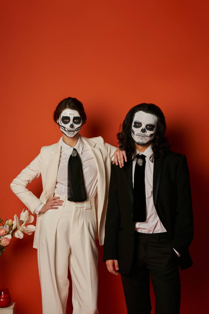 стильная пара в макияже черепа и элегантные костюмы позируют рядом с цветами и смотрят на камеру на красном - Фото, изображение