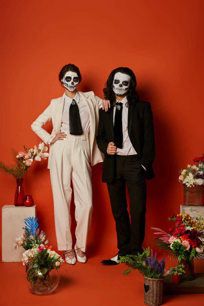 couple élégant dans le maquillage du crâne de sucre près de l'autel traditionnel dia de los muertos avec des fleurs sur rouge - Photo, image