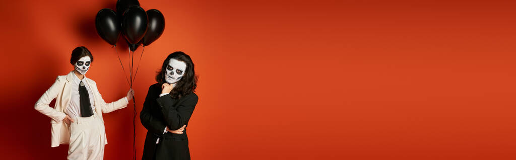 woman in sugar skull makeup and white suit with black balloons near spooky man on red, banner - Photo, Image