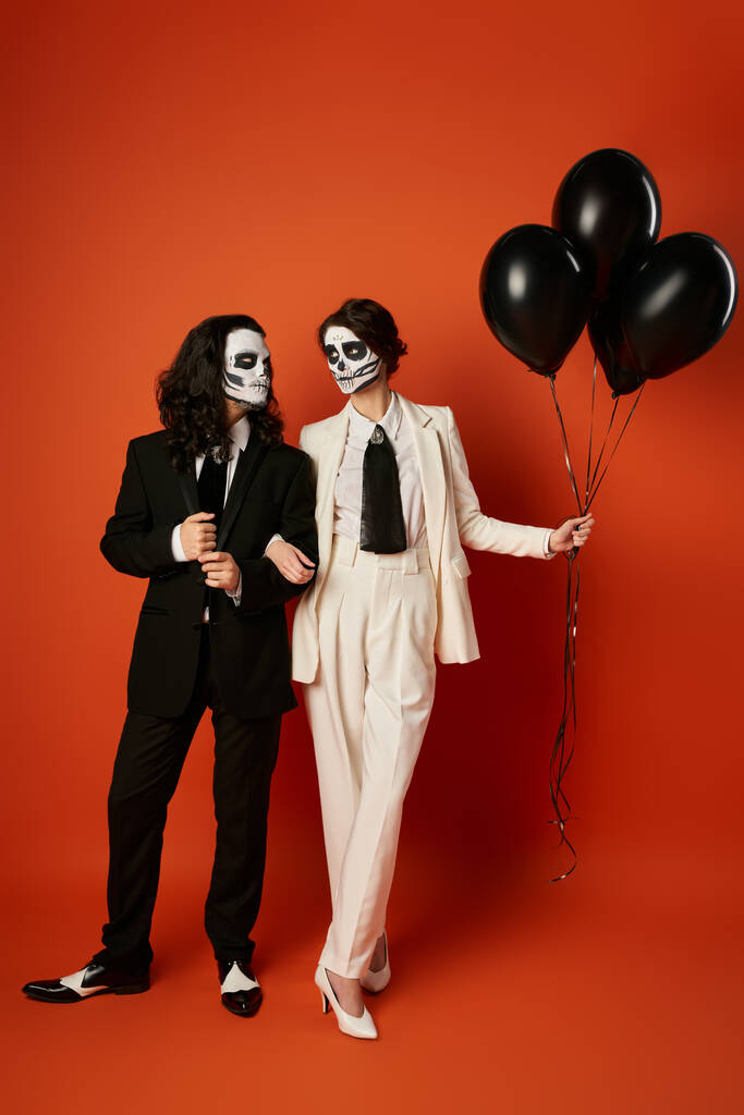 пара в котячому макіяжі і костюмах дивиться один на одного біля чорних кульок на червоному, День мертвих - Фото, зображення