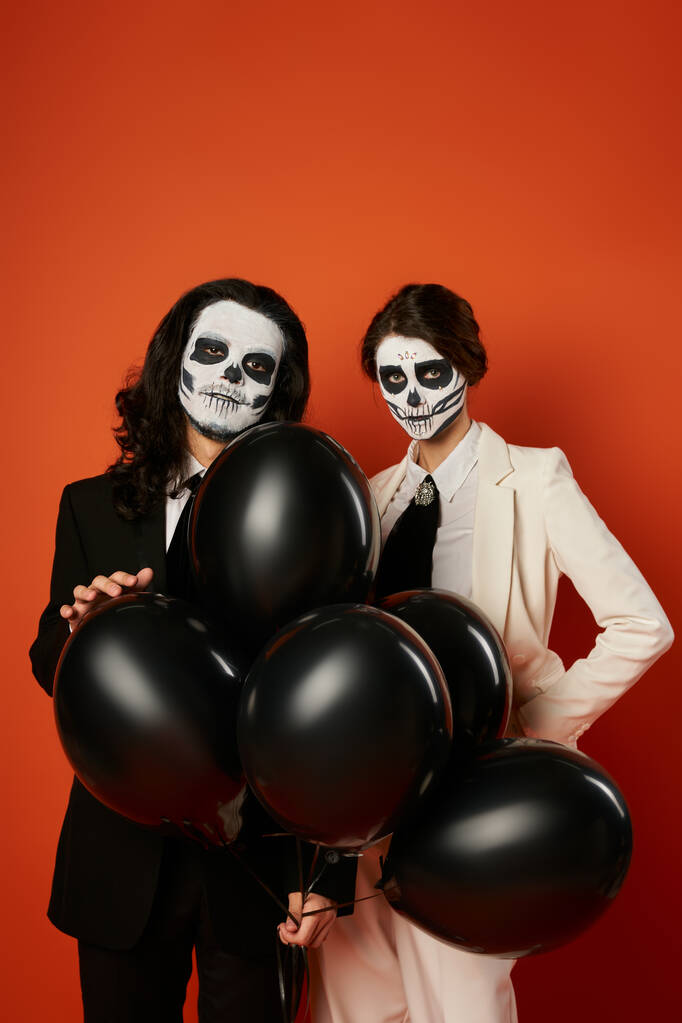 fiesta de día de los muertos, pareja espeluznante en maquillaje de cráneo mirando a la cámara cerca de globos negros sobre rojo - Foto, Imagen