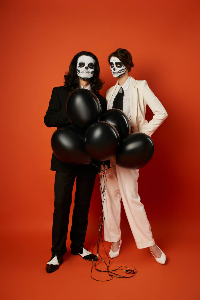 full length of couple in skull makeup and suits with black balloons on red, dia de los muertos party - Photo, Image