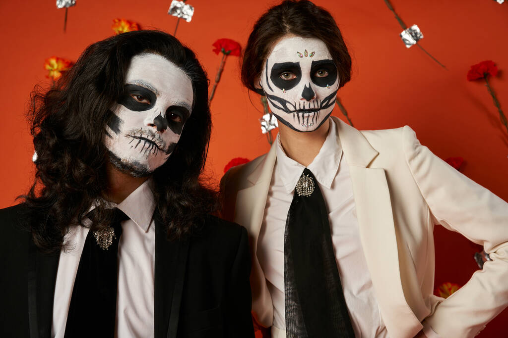 portrait of stylish couple in dia de los muertos makeup in red studio with carnation flowers decor - Photo, Image