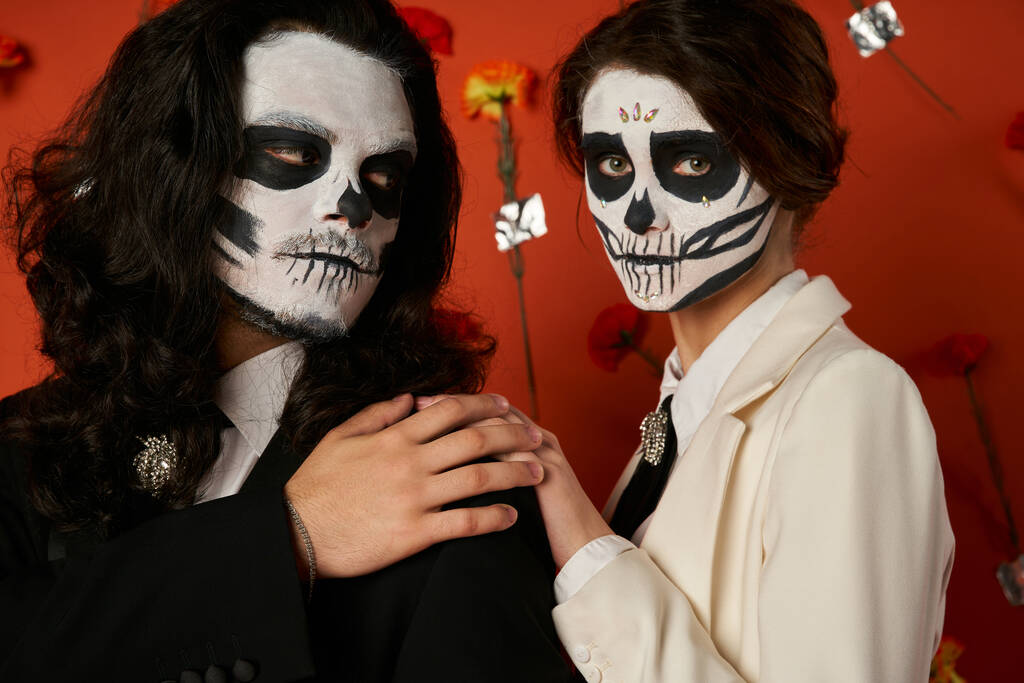 kobieta w dia de los muertos makijaż czaszki dotykając ramię mężczyzny na czerwonym tle z goździkami - Zdjęcie, obraz