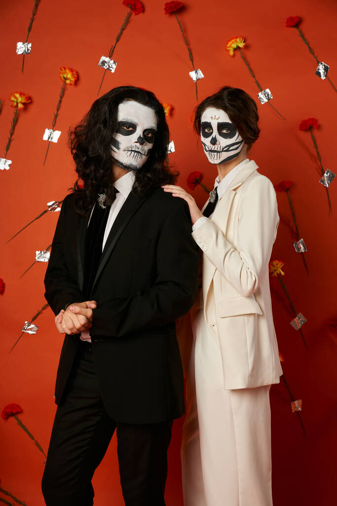 woman in dia de los muertos catrina makeup touching shoulder of man on red backdrop with carnations - Photo, Image