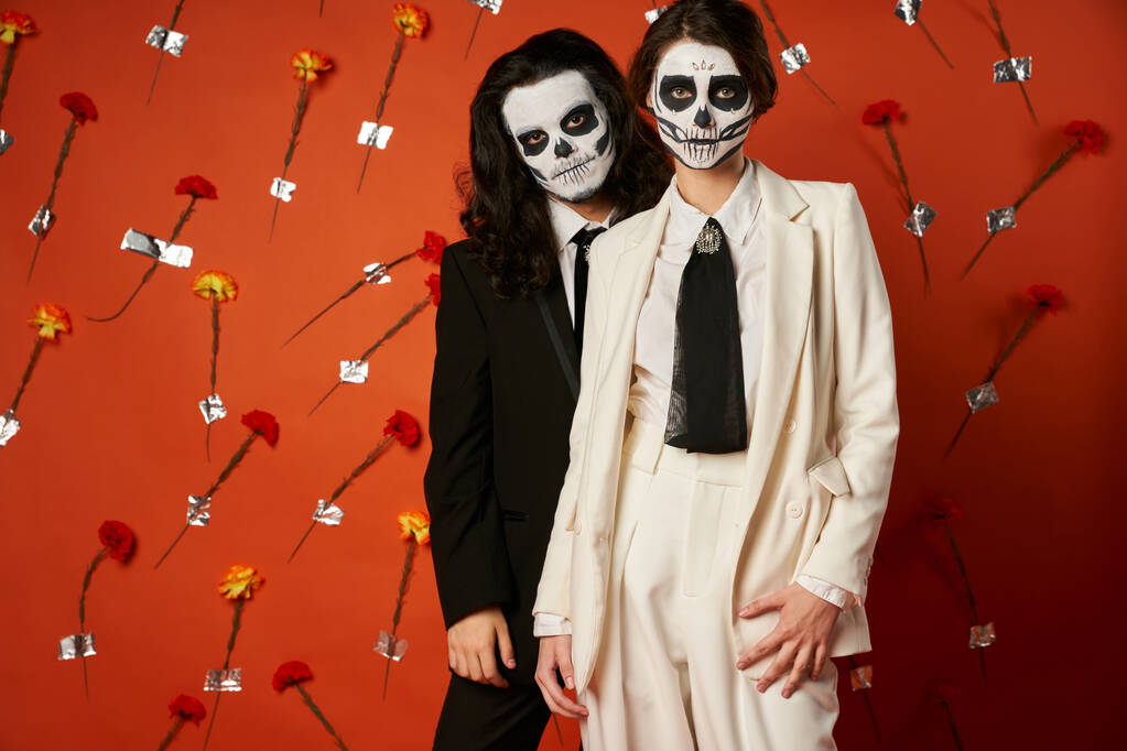 couple in dia de los muertos catrina makeup and suits looking at camera on red backdrop with flowers - Photo, Image