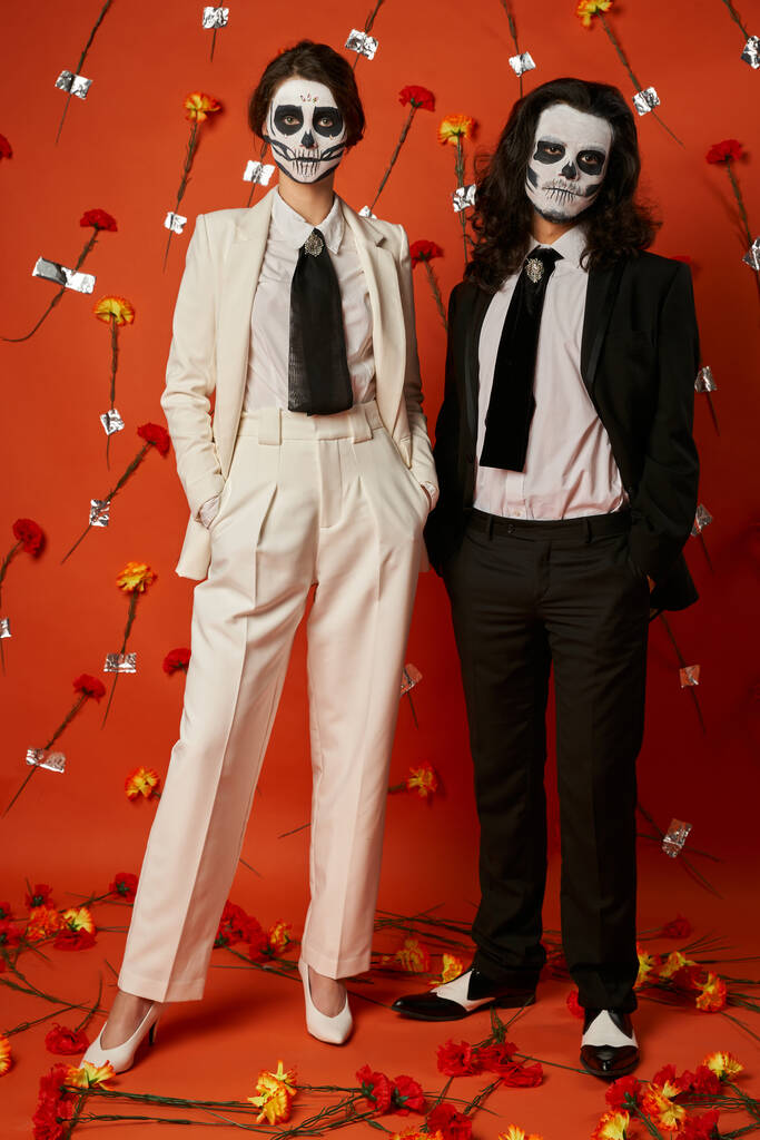 paar in suiker schedel make-up en trendy outfit poseren met de handen in zakken van rode bloemen achtergrond - Foto, afbeelding