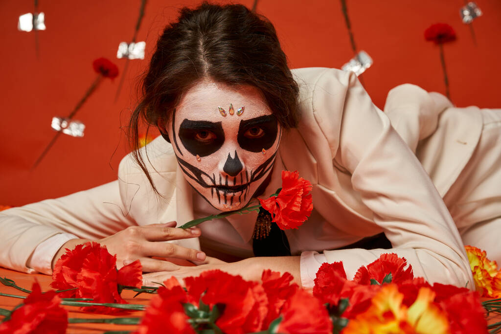 femme élégante dans le maquillage effrayant catrina couché et regardant la caméra près des fleurs sur fond rouge - Photo, image