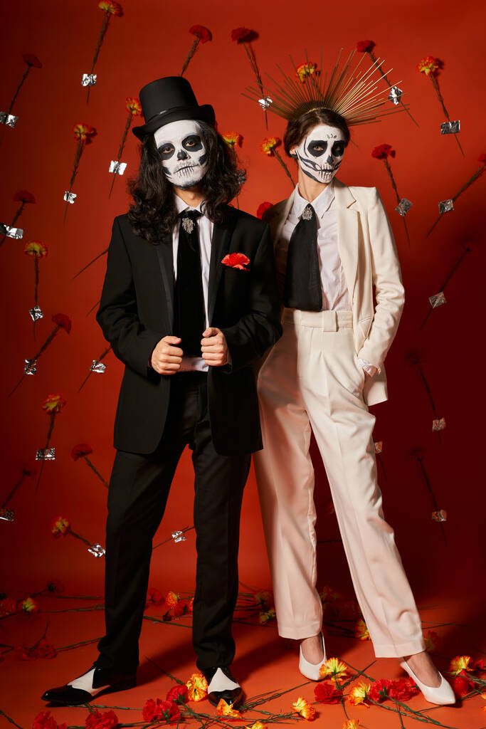 full length of elegant couple in dia de los muertos makeup on red backdrop with floral decor - Photo, Image