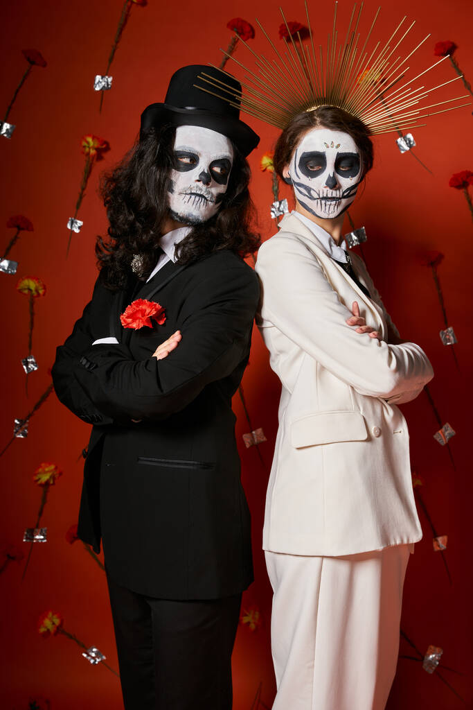 элегантная пара в сахарном черепе макияж стоя спиной к спине со сложенными руками на красном цветочном фоне - Фото, изображение