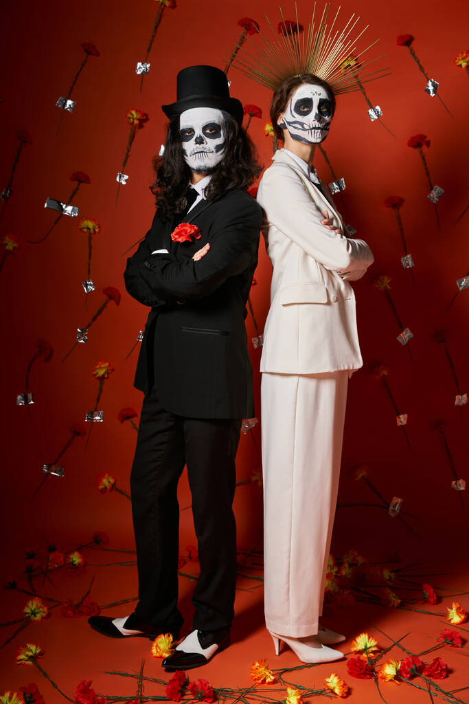 trendy couple in catrina makeup standing back to back with folded arms on red backdrop with flowers - Photo, Image
