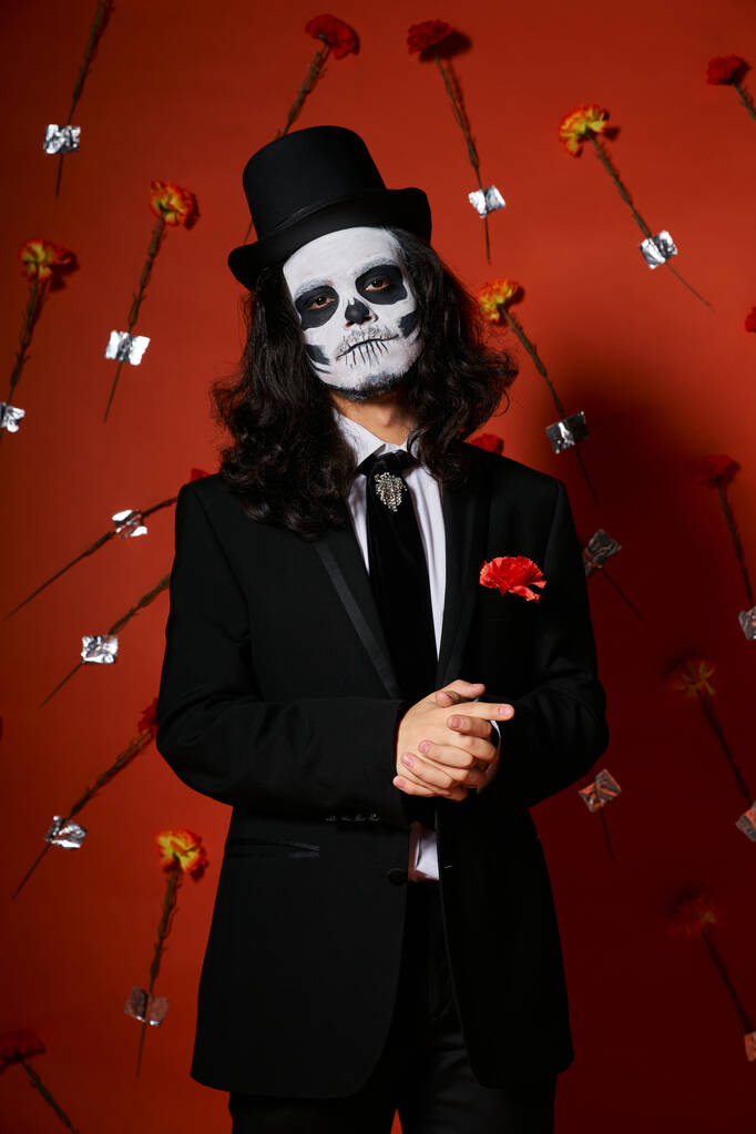 man in skull makeup and festive attire looking at camera on red floral backdrop, dia de los muertos - Photo, Image