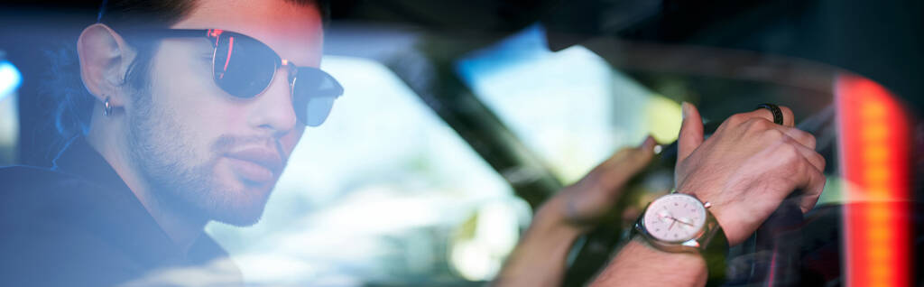 pohledný mladý muž ve slunečních brýlích pózující na volantu svého auta s náramkovými hodinkami, prapor - Fotografie, Obrázek