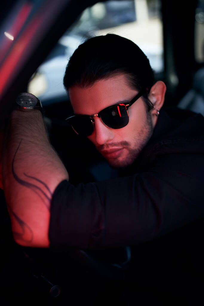 sexy mladý řidič s tetováním a slunečními brýlemi pózovat na volantu auta při pohledu do kamery - Fotografie, Obrázek