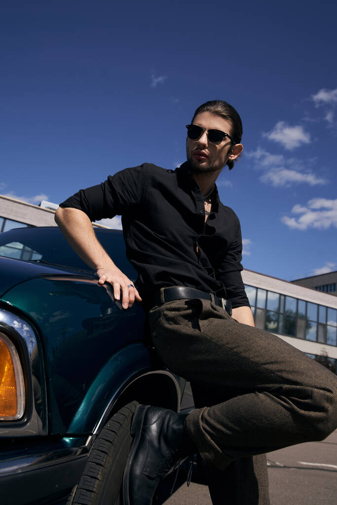 homme barbu élégant avec tatouage et lunettes de soleil en tenue noire debout à côté de sa voiture, chauffeur sexy - Photo, image