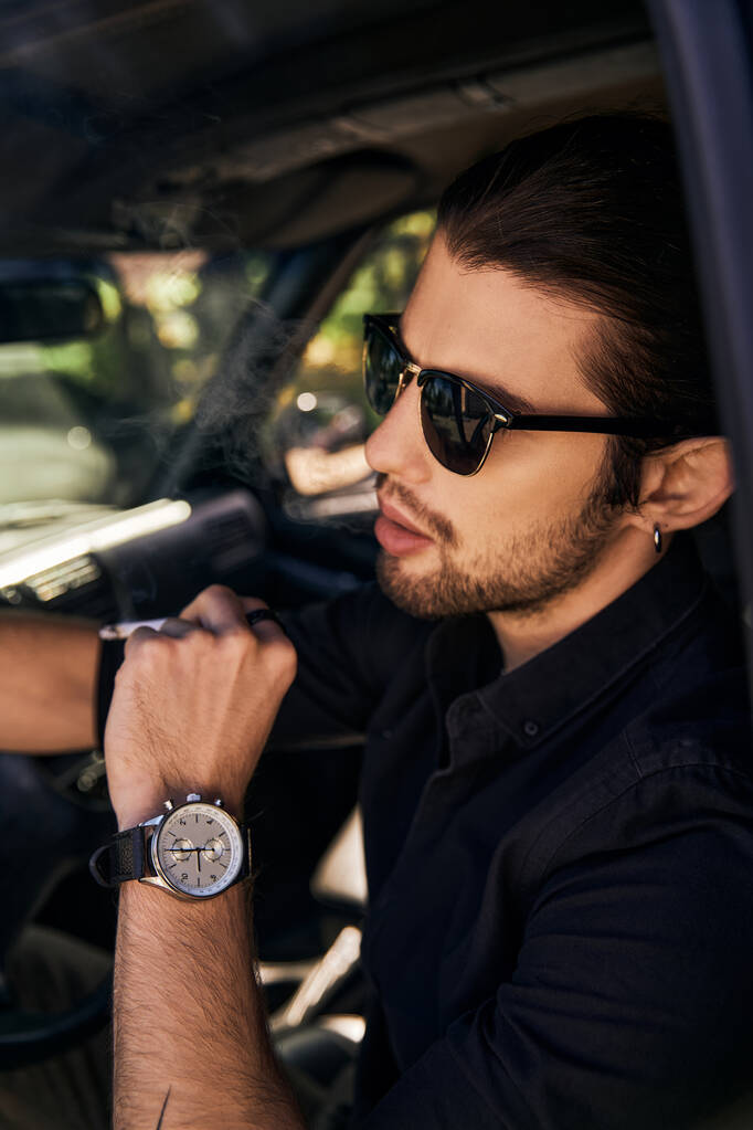 εμφανίσιμος νεαρός άνδρας με γυαλιά ηλίου και ρολόι χειρός κάθεται στο αυτοκίνητό του και ποζάρει στο προφίλ - Φωτογραφία, εικόνα