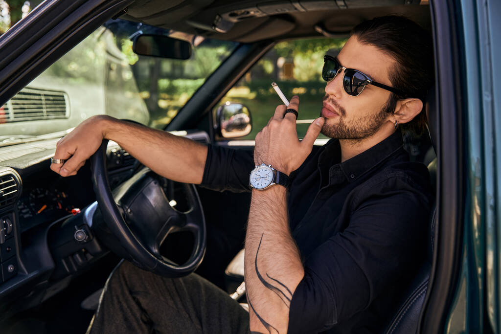 гарний виглядає бородатий чоловік з хвостом і сонцезахисними окулярами палить сигарету, позуючи в його машині - Фото, зображення
