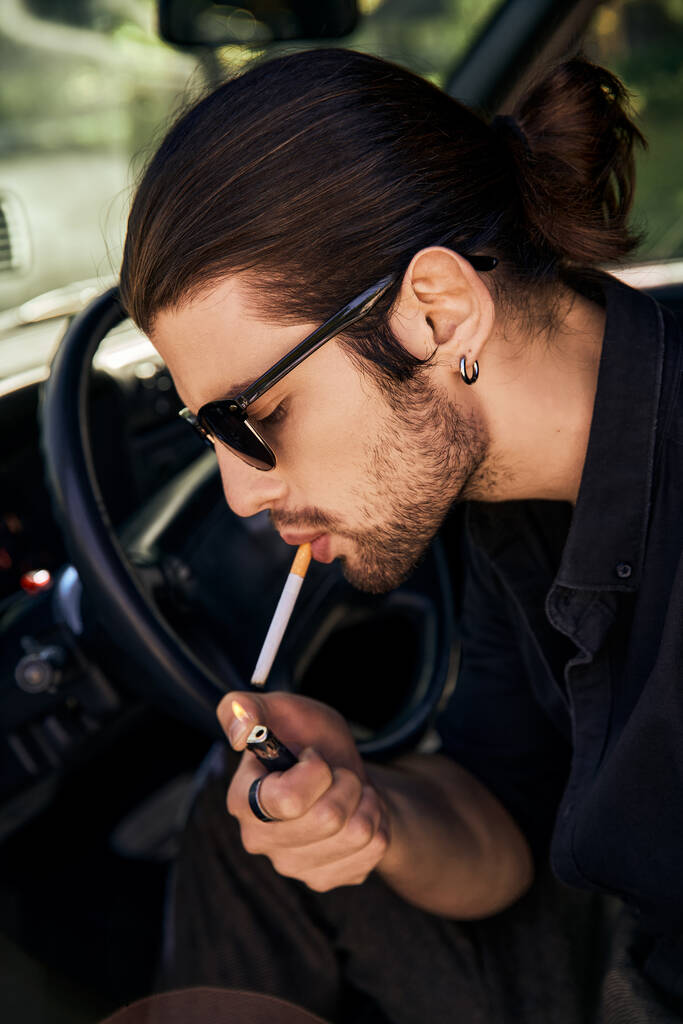 handsome sexy male model with earring and sunglasses lighting up his cigarette in car, sexy driver - Фото, изображение
