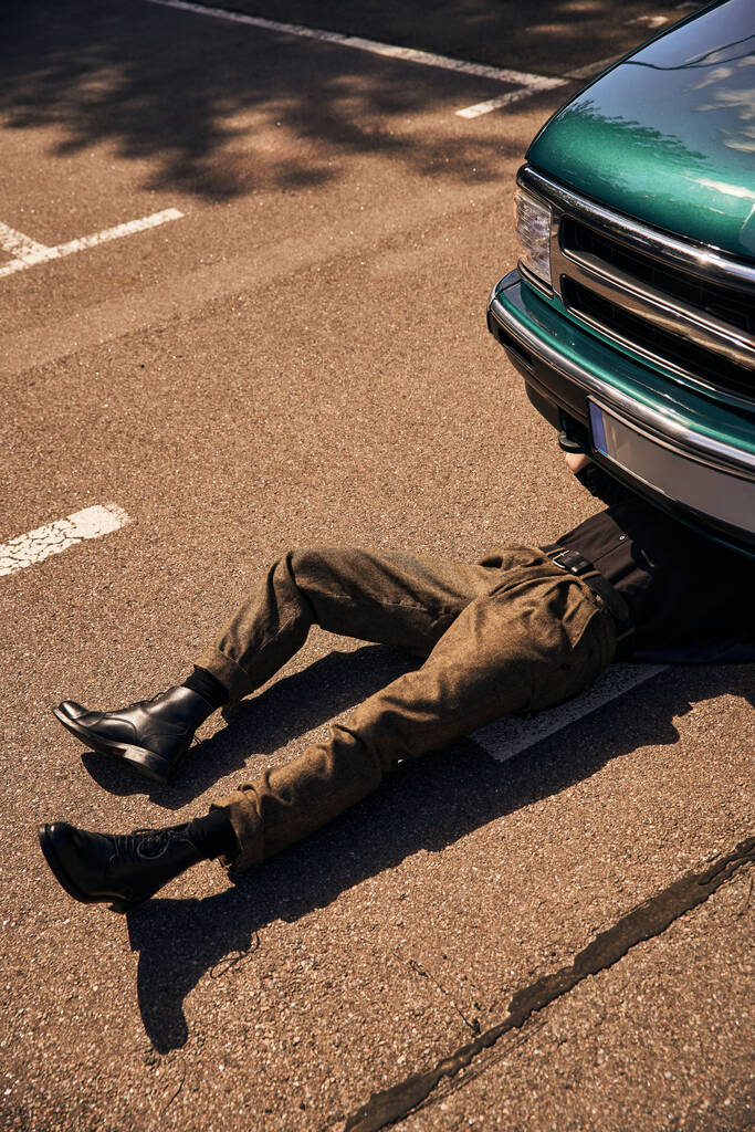 νεαρό κομψό ανδρικό μοντέλο σε μαύρο μοντέρνο αστικό ντύσιμο που βρίσκεται κάτω από το αυτοκίνητό του και την επισκευή - Φωτογραφία, εικόνα