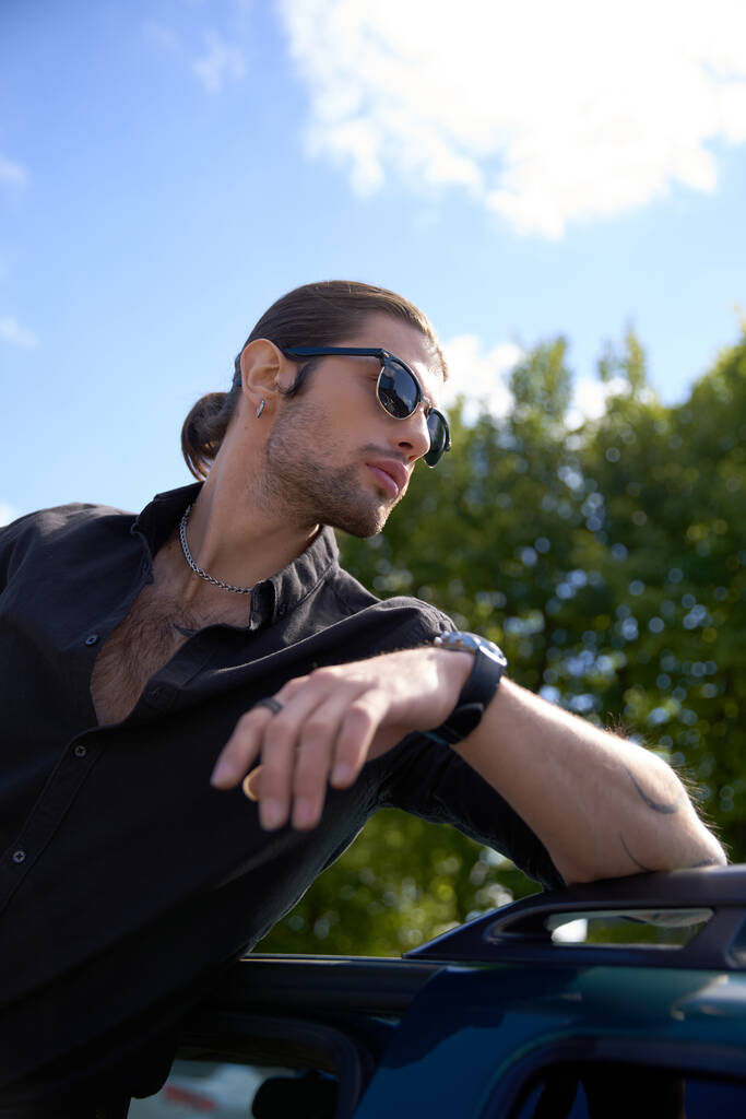 verticaal schot van sexy jonge bestuurder met accessoires in zwart outfit poseren met elleboog op zijn auto - Foto, afbeelding